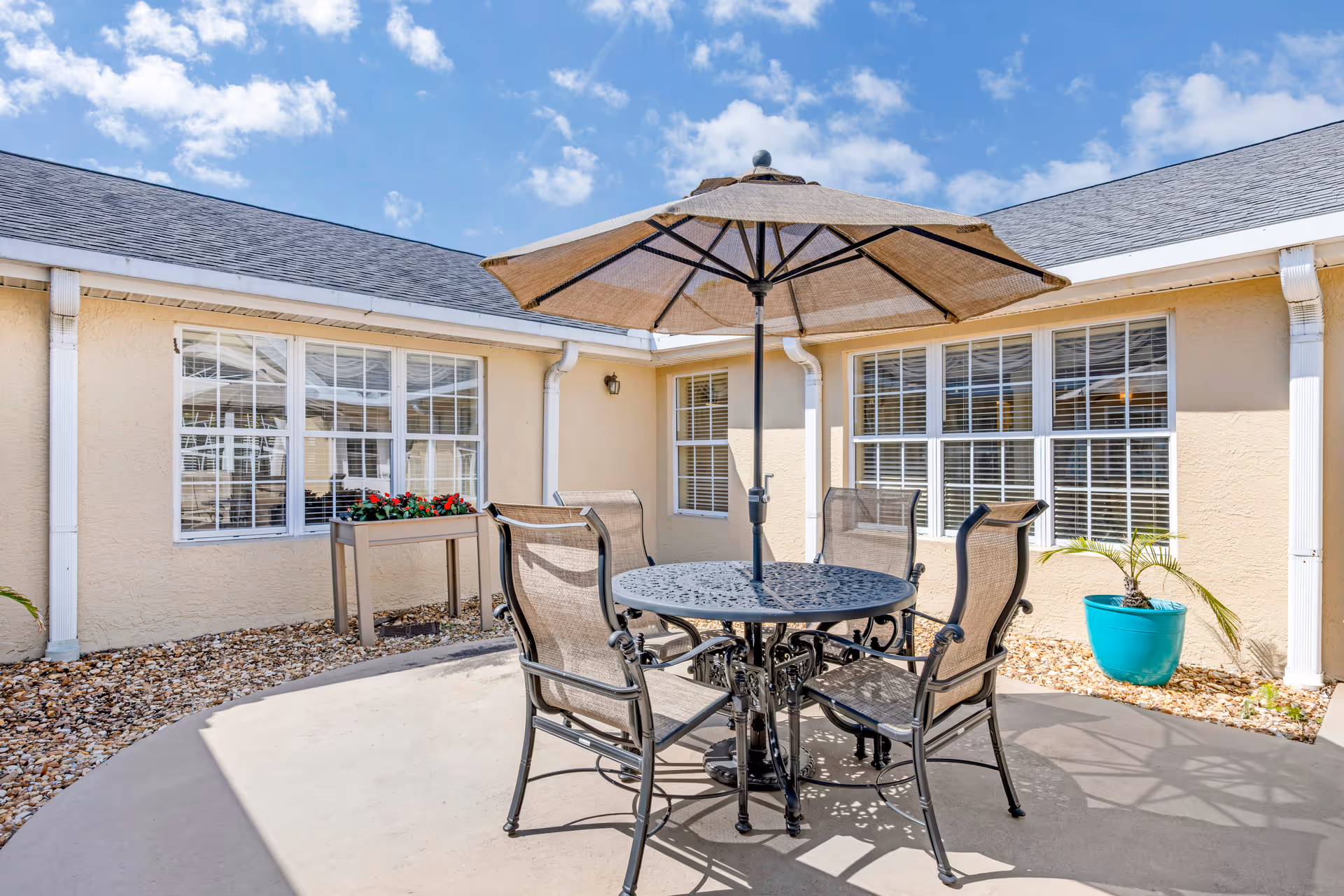 Outdoor patio area with a round metal table and four mesh chairs under a large beige umbrella. The patio is surrounded by a beige building with multiple windows and white trim. There is a small planter with red flowers and a turquoise pot with a small palm plant on the gravel-covered ground around the patio.