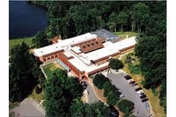 Aerial view of a large, single-story building surrounded by trees and greenery, with a parking lot on one side and a body of water nearby. The building has a white roof and brick exterior walls.