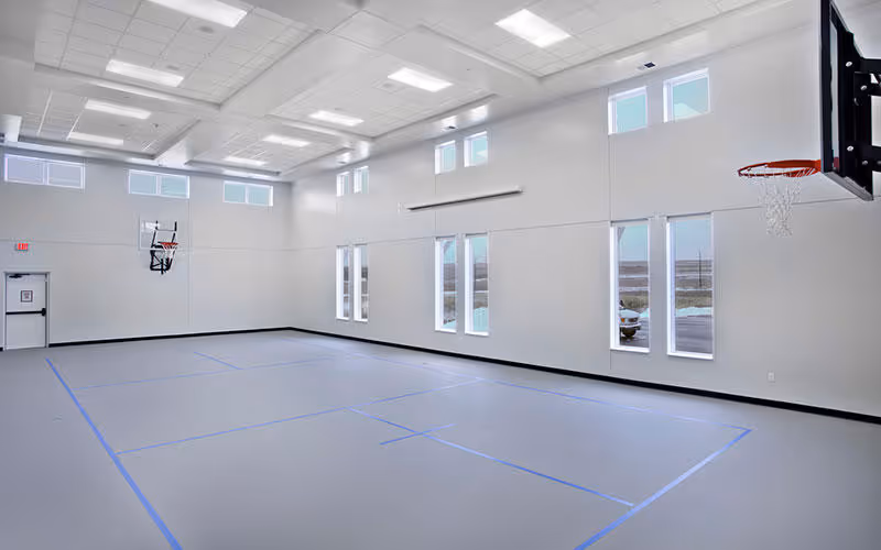 An empty indoor basketball court with light gray flooring marked with blue lines, two basketball hoops mounted on opposite walls, high windows letting in natural light, and a white ceiling with recessed lighting panels.