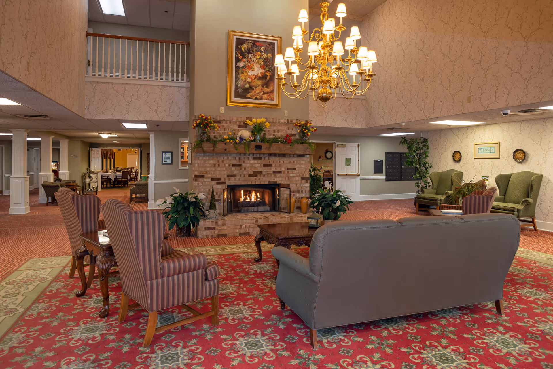 A cozy senior living facility common area with a brick fireplace in the center, decorated with flowers and a painting above it. The room features a large ornate chandelier, patterned red carpet, striped armchairs, a gray sofa, green armchairs, and a wooden coffee table. The walls have a light floral wallpaper and there is a balcony railing visible on the upper level.