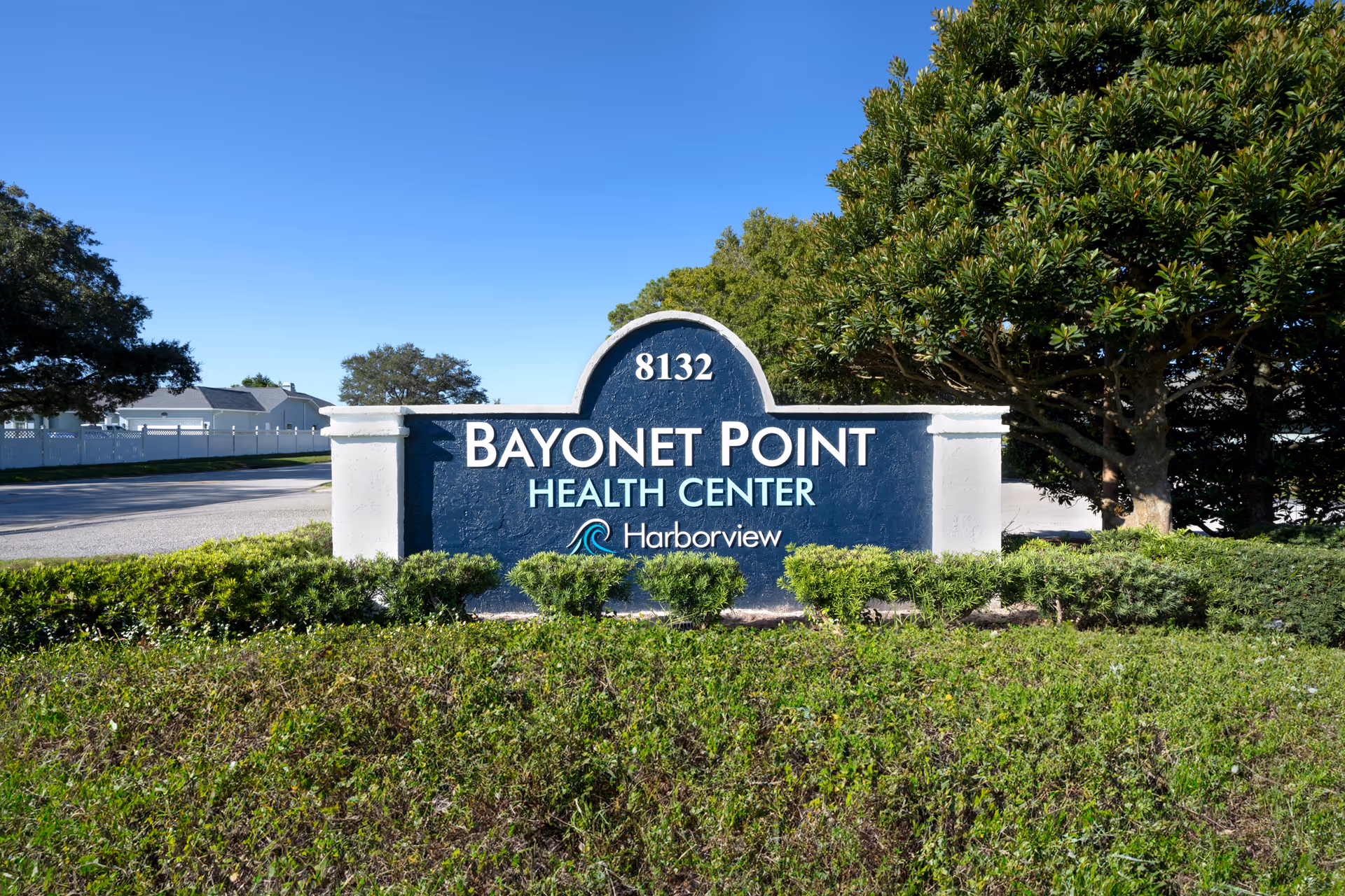 Outdoor view of a blue and white sign for Bayonet Point Health Center with the number 8132 and the Harborview logo, surrounded by green bushes and trees under a clear blue sky.