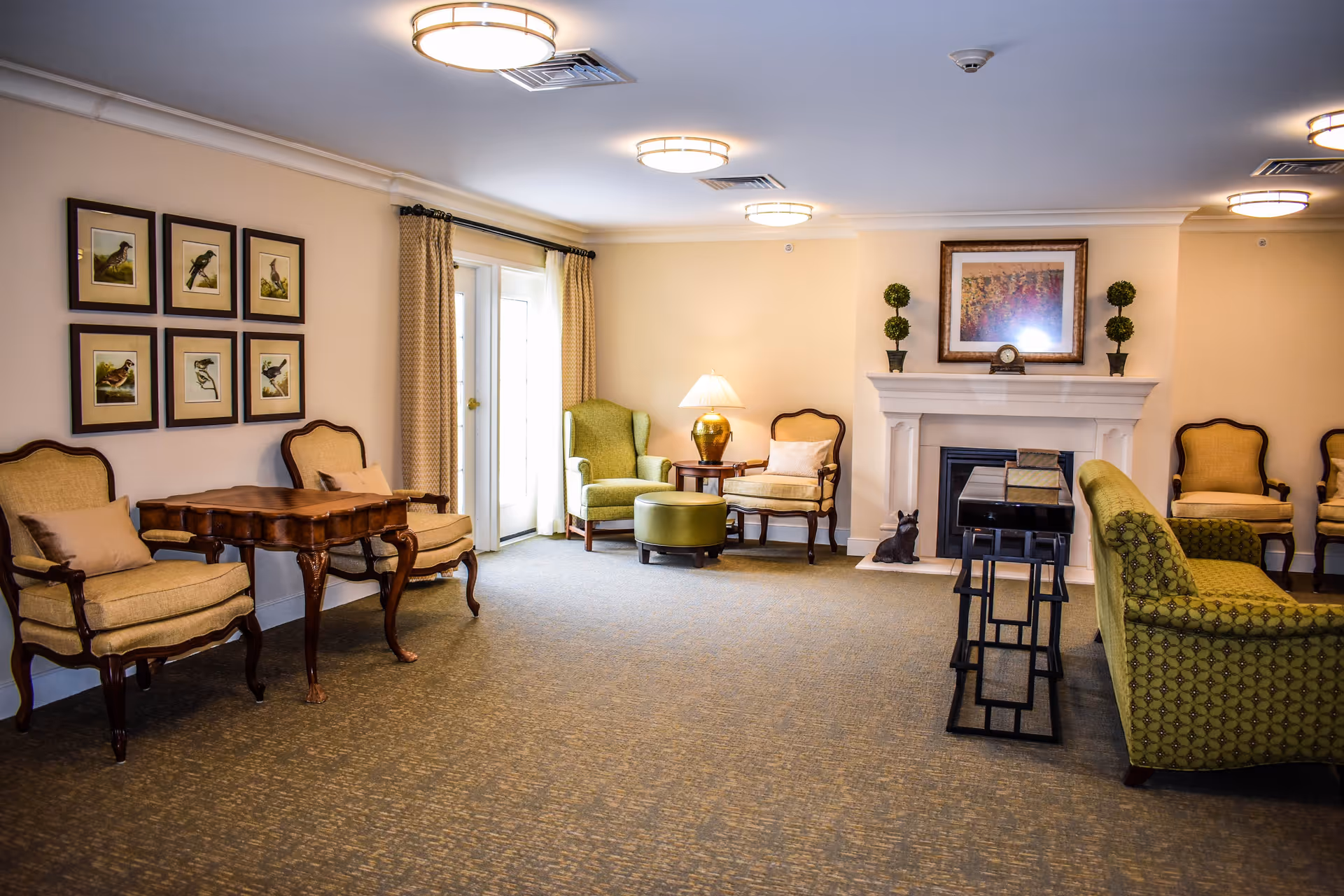A cozy living room area with beige walls and carpeted floor. The room features several upholstered chairs, a green patterned sofa, a wooden table, and a green armchair with an ottoman near a set of glass doors with curtains. There is a white fireplace with decorative plants and a framed picture above it. The room is well-lit with ceiling lights and a table lamp.