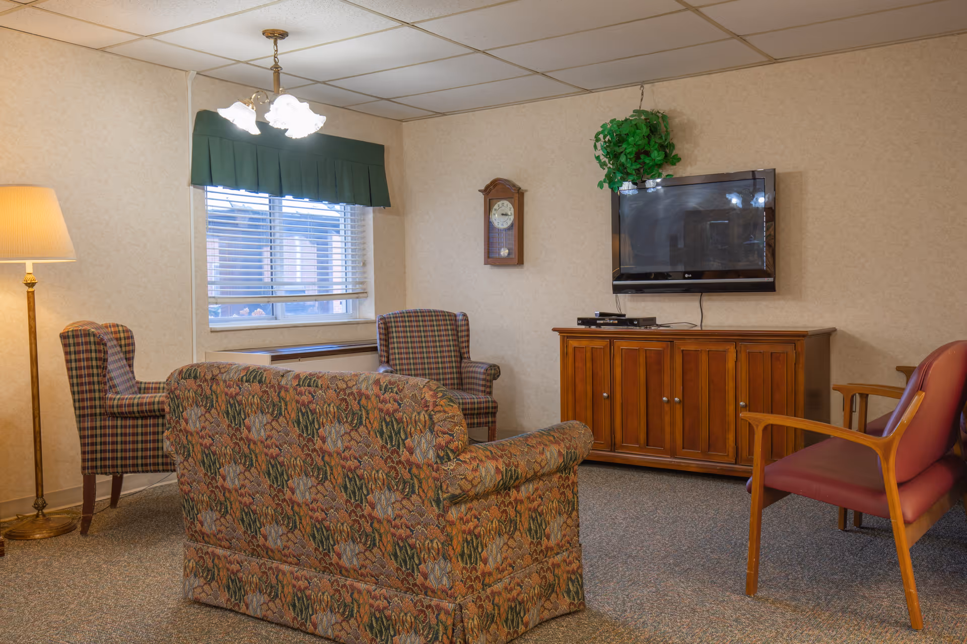 Cozy living room with patterned sofa and armchairs, a wall-mounted TV above a wooden cabinet, and a window with a green valance.