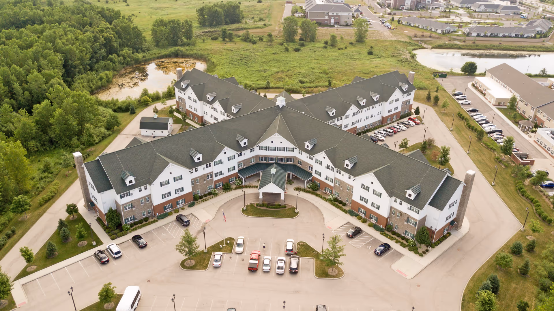 Aerial view of Deer Path of Huntley senior living facility showing a large, multi-wing building with a green roof and white and brick exterior. The building is surrounded by parking lots with cars, green trees, grassy areas, and a small pond in the background.