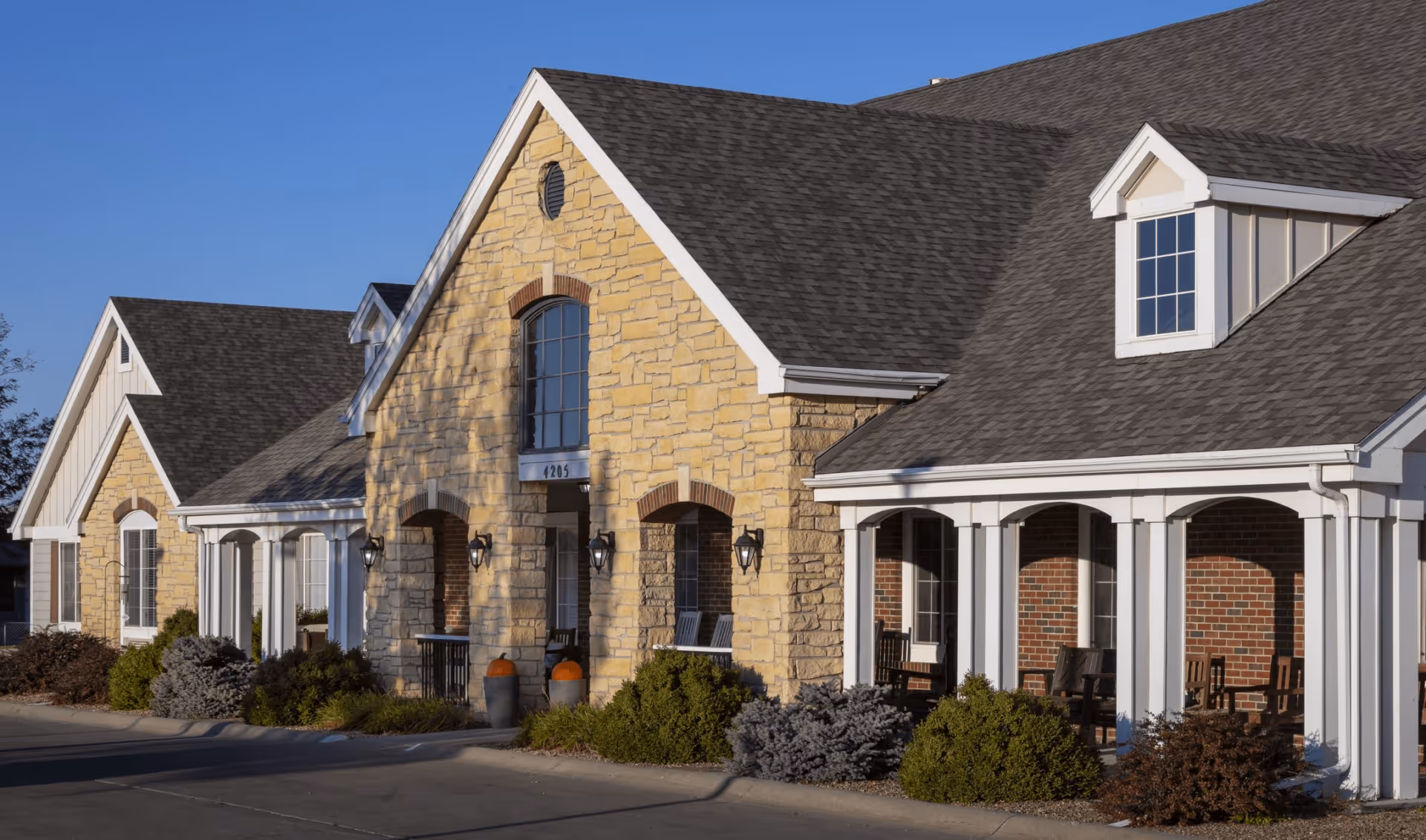 Front exterior of a single-story stone and brick assisted living building with a columned porch, arched entrances, and landscaped shrubs.