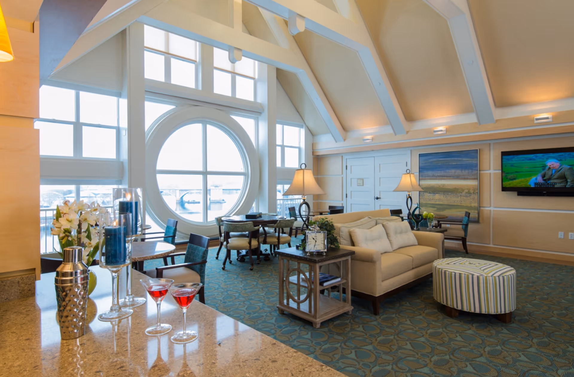 Bright communal lounge with vaulted ceiling and a large round window overlooking the waterfront, furnished with sofas, tables and a TV.