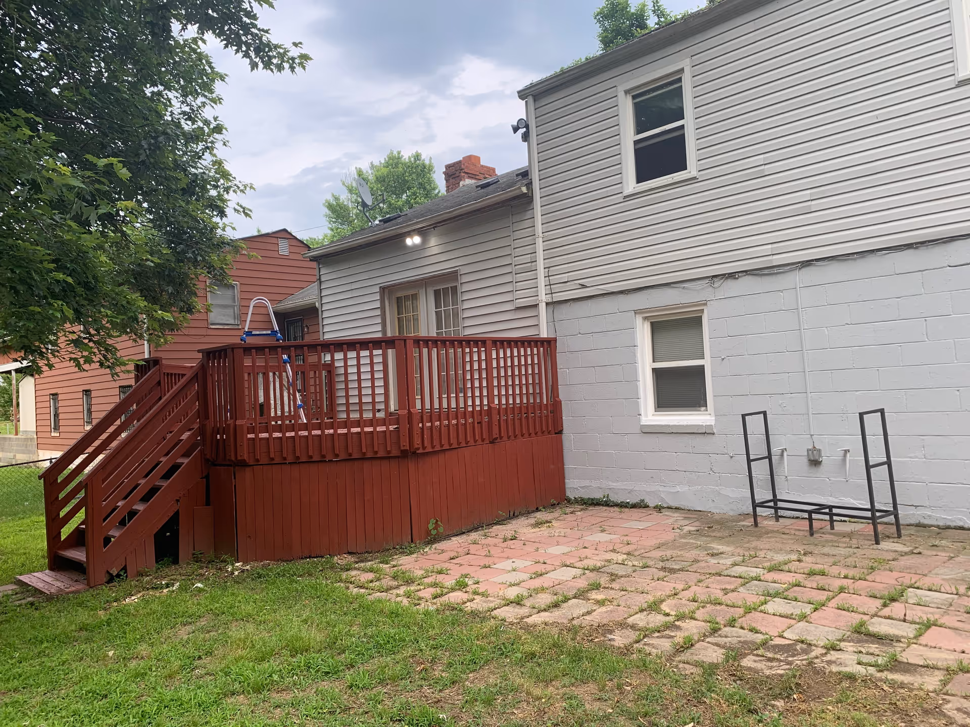 Backyard area of a residential building with a red wooden deck and stairs, a paved patio with some grass growing between the stones, and a tree on the left side. The building has light gray siding and two windows visible.