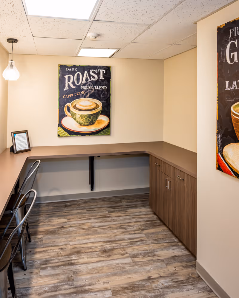 A small room with a long L-shaped counter along two walls, two metal chairs, wood-look flooring, beige walls, and a ceiling with recessed lighting and a hanging light fixture. A framed coffee-themed artwork featuring a cup of cappuccino and the words 'Dark Roast House Blend Cappuccino' hangs on the wall.