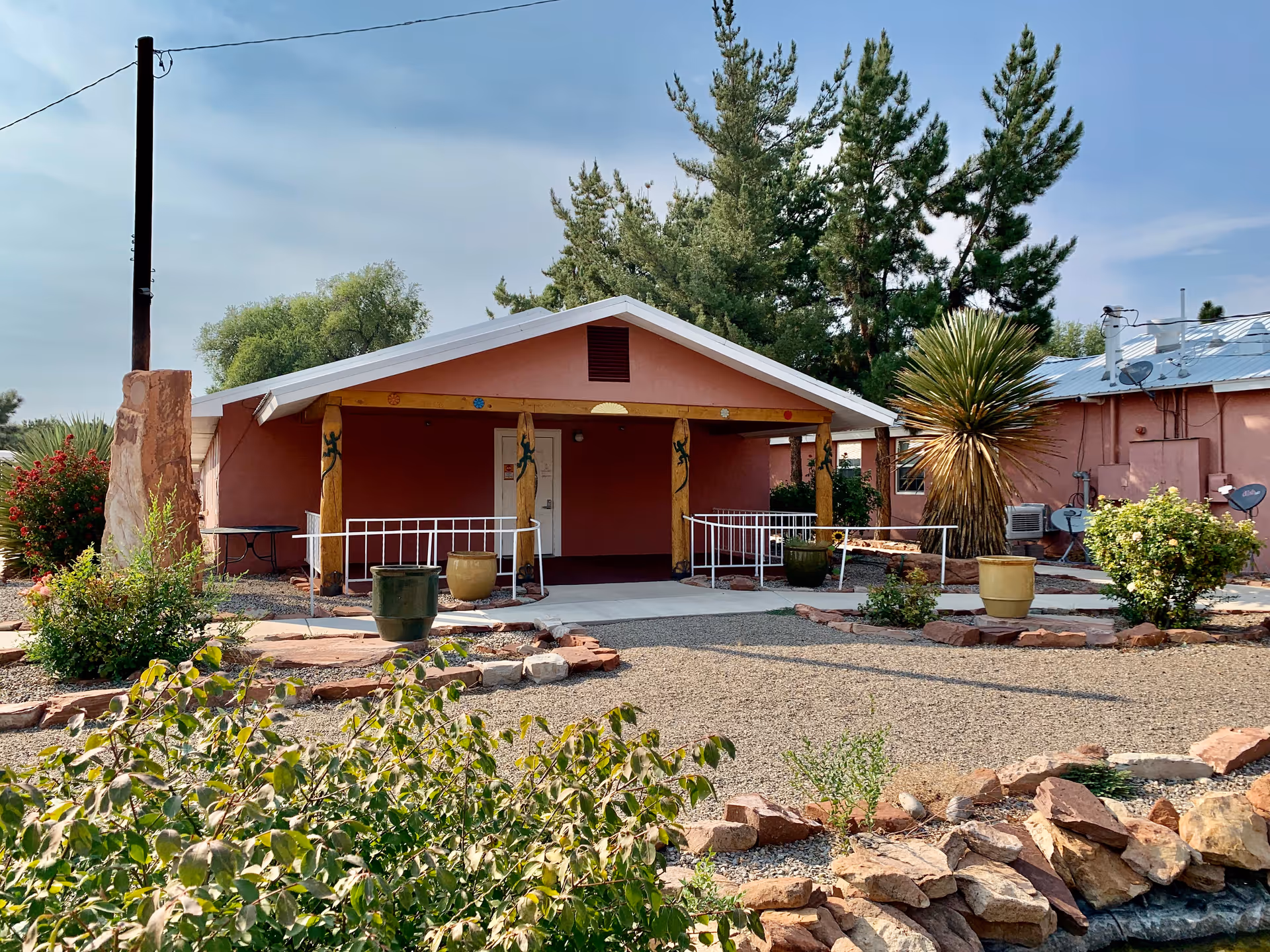Exterior view of a single-story building with a covered porch supported by wooden posts decorated with lizard designs. The building is painted in a reddish color and surrounded by a landscaped garden with various plants, shrubs, and stone borders under a partly cloudy sky.