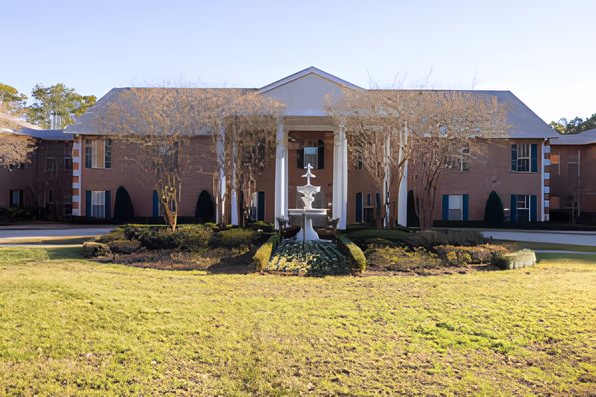 Front exterior view of a two-story brick building with white columns and a central fountain surrounded by landscaped bushes and grass under a clear sky.