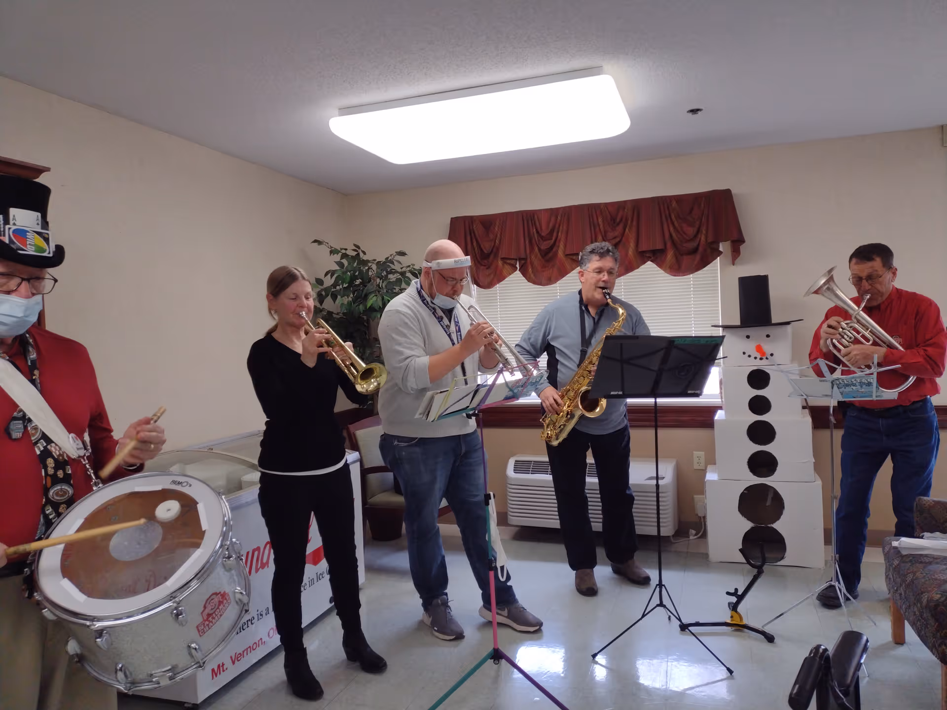 A group of five musicians playing instruments indoors. One person is playing a drum, two are playing trumpets, one is playing a saxophone, and one is playing a euphonium. They are standing in a room with beige walls, a window with red curtains, a potted plant, and a large cardboard snowman decoration.