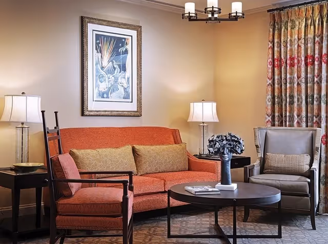 Cozy living room with an orange sofa, two armchairs, a round coffee table, lamps, framed artwork, and patterned curtains.