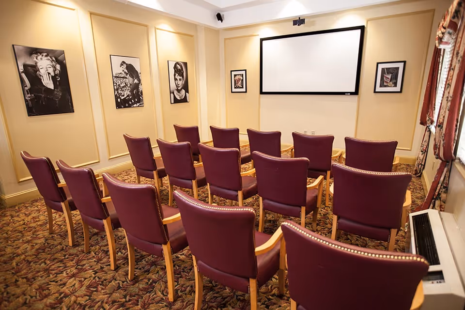 A small theater or presentation room with three rows of maroon chairs facing a large white projection screen. The walls are decorated with black and white framed photographs of classic movie stars. The room has patterned carpet and a window with floral curtains on the right side.