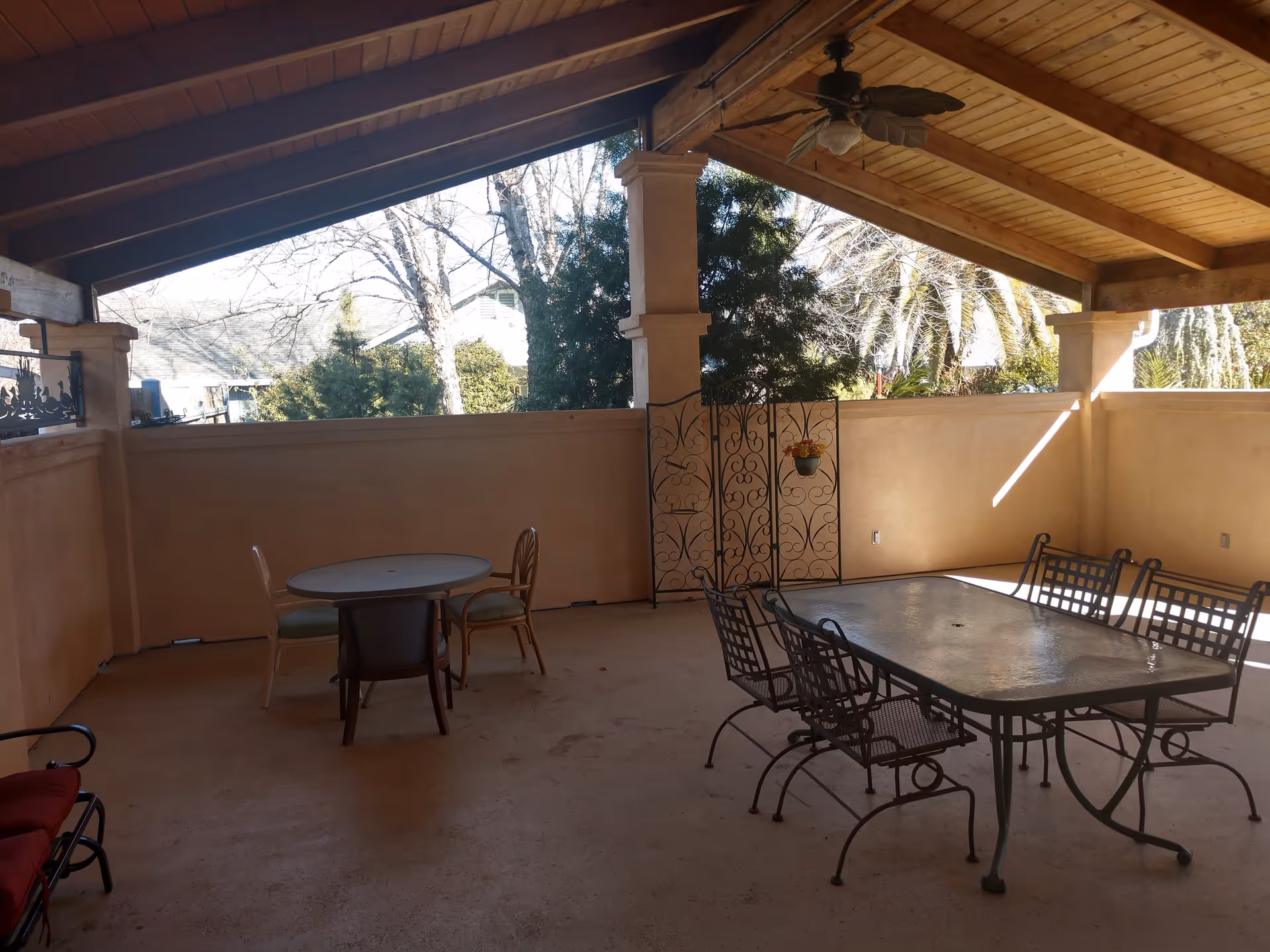 Covered outdoor patio with a wooden vaulted ceiling and fan, a metal dining table with chairs, a small round table and chairs by a half wall overlooking trees.