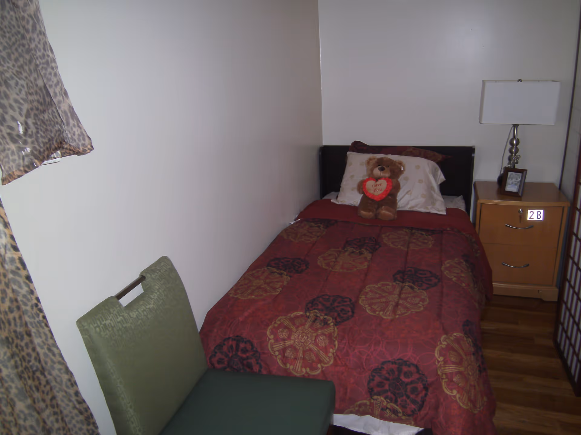 A small bedroom with a single bed covered in a red and gold patterned comforter. A teddy bear holding a heart that says 'Love You' is placed on the pillow. Next to the bed is a wooden nightstand with a lamp and a framed photo. A green chair is positioned near the bed, and a leopard print curtain is partially visible on the left side.