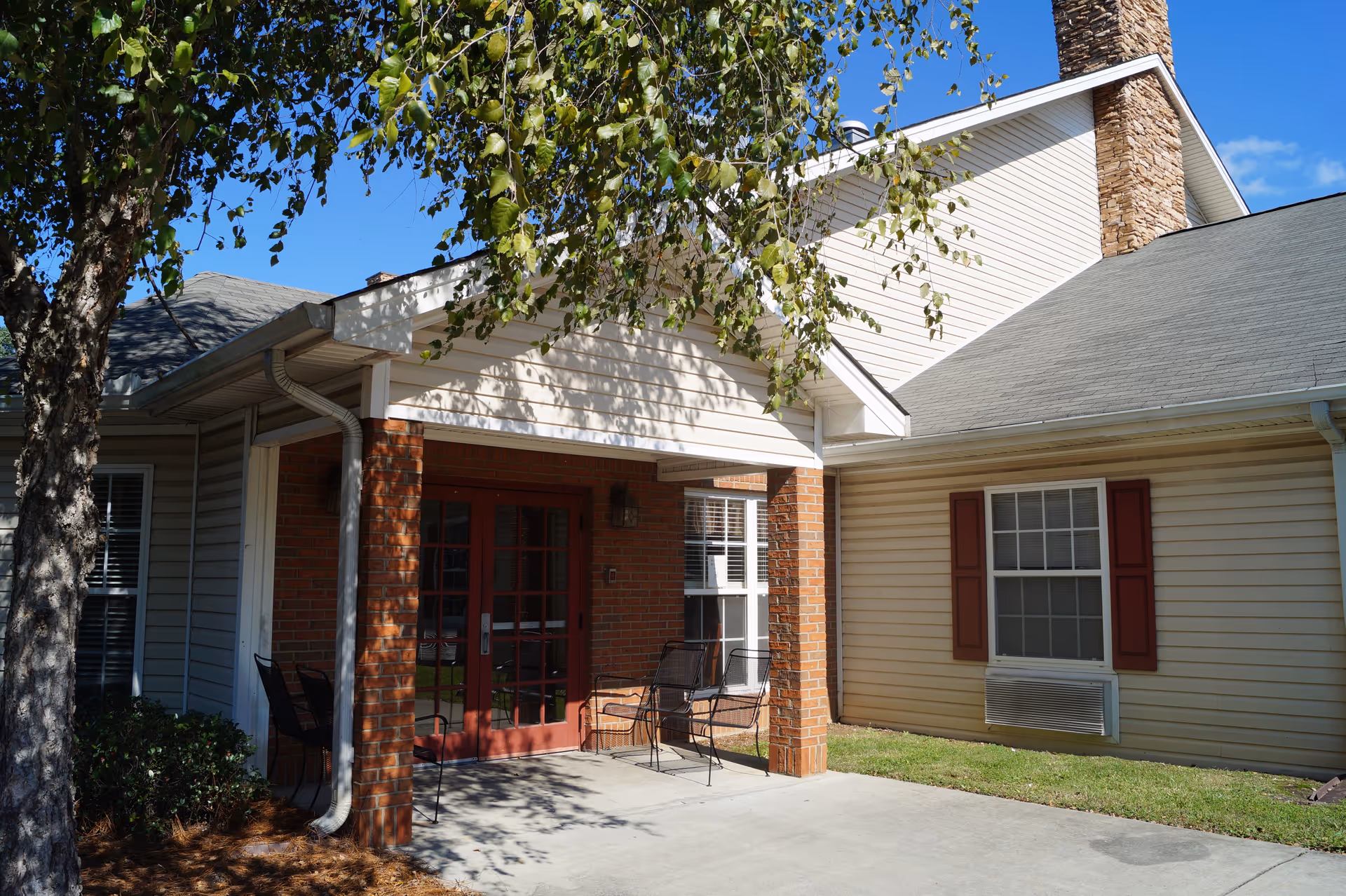 Exterior view of a building entrance with a covered porch supported by brick columns. There are two black metal chairs on the porch, a tree with green leaves casting shadows, and a window with red shutters and an air conditioning unit below it. The sky is clear and blue.