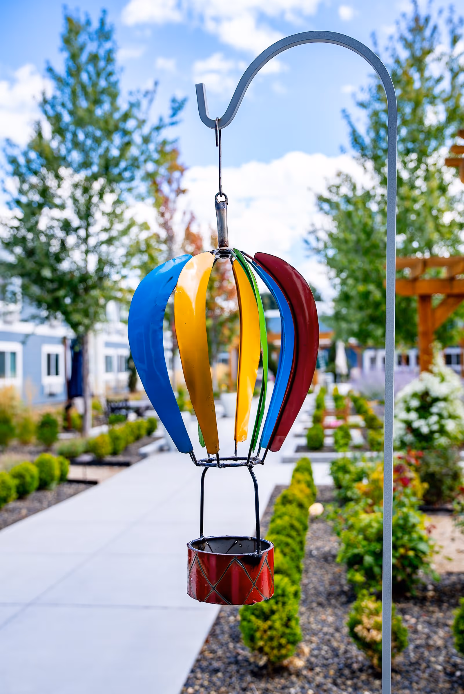 Colorful metal hot air balloon garden decoration hanging from a curved metal hook along a paved walkway with green shrubs and trees in the background at Heatherwood Senior Living.