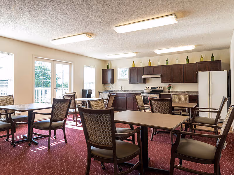 A bright dining area with multiple tables and chairs arranged on a red carpet. The room features a kitchen area with dark wood cabinets, a white refrigerator, stove, and a sink. Several decorative bottles are placed on top of the cabinets. Large windows and a glass door allow natural light to fill the space.