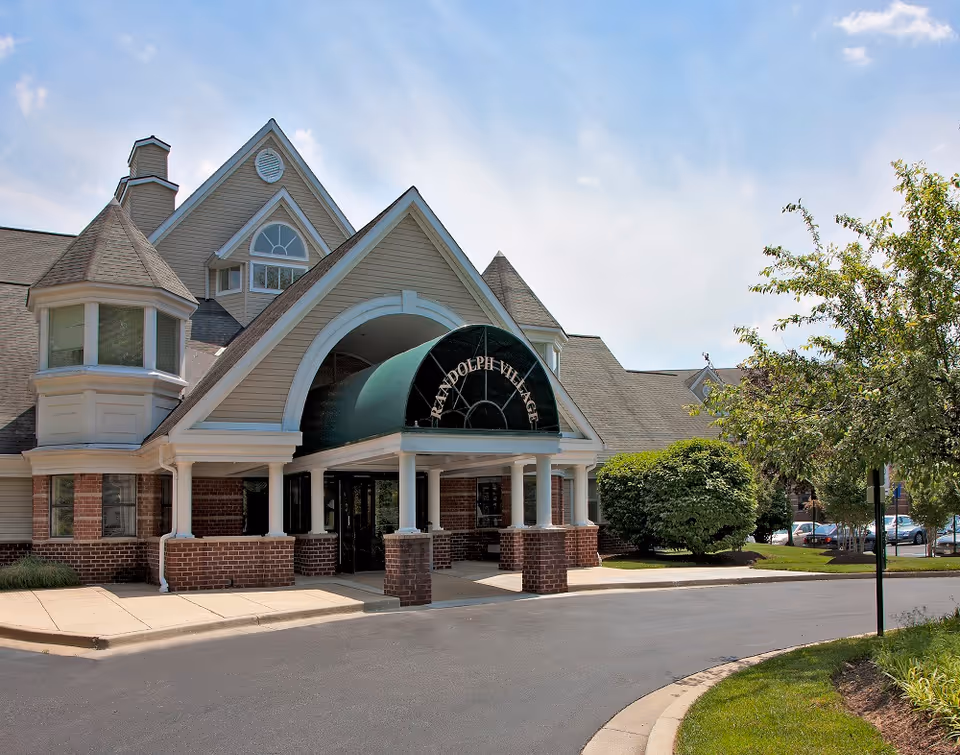 Front entrance of Randolph Village showing a covered porte-cochère with brick pillars, arched canopy, and surrounding landscaping.