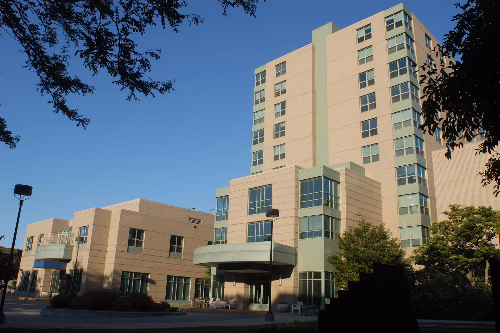 Exterior view of a multi-story senior living facility building with large windows, beige and green facade, surrounded by trees and outdoor seating under a clear blue sky.