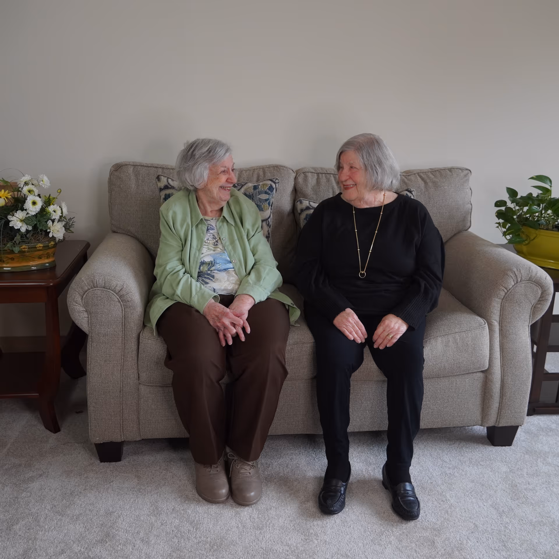 Two elderly women sitting on a beige couch in a living room, smiling and looking at each other. There are floral arrangements on wooden side tables on either side of the couch.