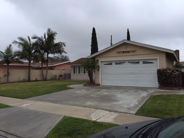 Front exterior view of a single-story house with a two-car garage, a concrete driveway, green lawn, palm trees, and overcast sky.