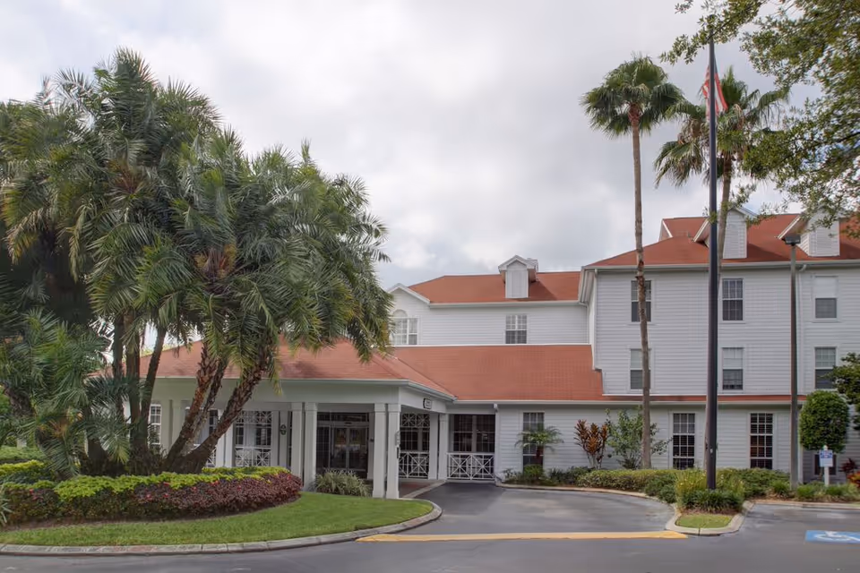 Exterior view of a senior living facility named Sodalis of Largo, featuring a white multi-story building with a red roof, surrounded by palm trees and landscaped greenery. There is a driveway leading to the entrance with a covered drop-off area and an American flag on a flagpole.