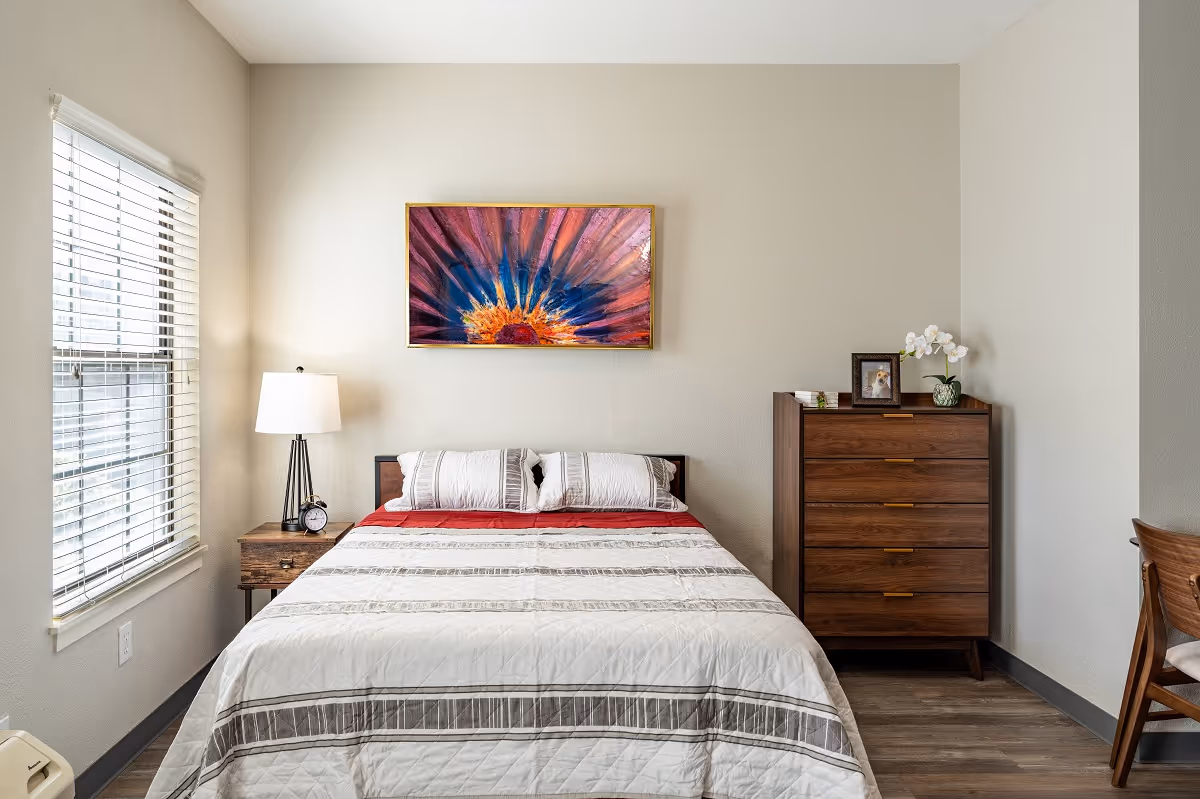 A tidy bedroom with a bed centered under a colorful painting, a bedside table and lamp by a window, and a wooden chest of drawers.