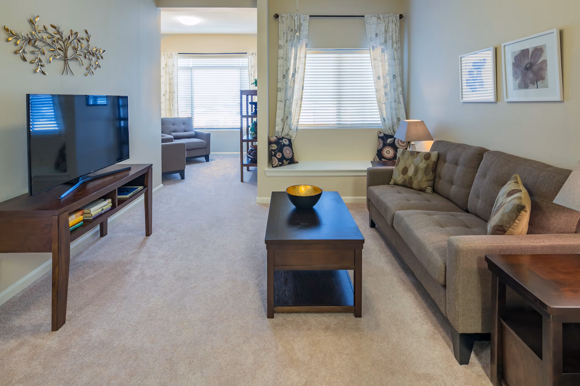 A cozy living room with a brown sofa adorned with patterned pillows, a dark wooden coffee table with a decorative bowl, and a matching TV stand holding a flat-screen television. The room has beige carpet flooring and light-colored walls with two framed artworks. A window with patterned curtains and a built-in window seat with cushions is visible, along with an adjacent sitting area featuring a gray armchair and a shelving unit.