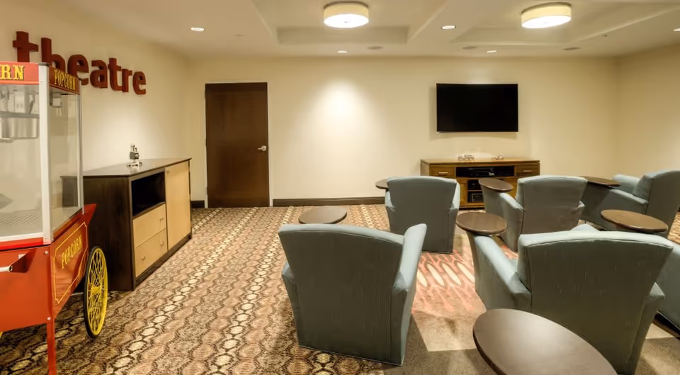 A small theater room with several blue upholstered chairs arranged in rows facing a wall-mounted flat screen TV. There is a wooden media console beneath the TV and a popcorn machine on the left side of the room. The walls are light-colored and the carpet has a patterned design.
