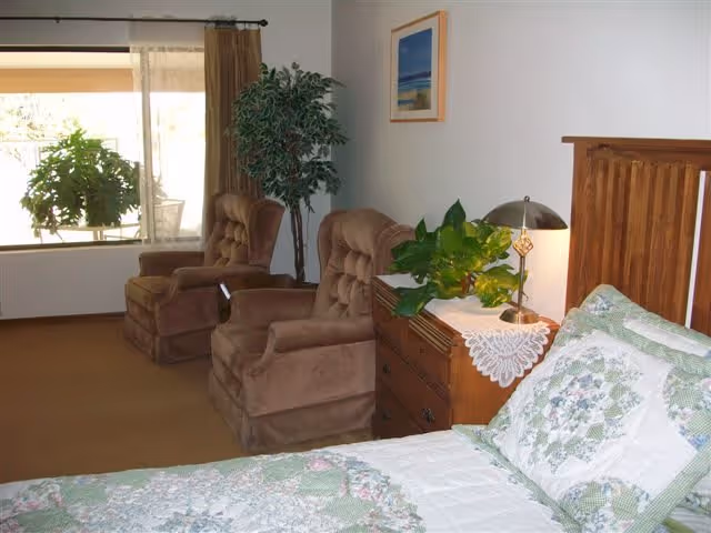 A cozy bedroom with a bed covered in a floral quilt and matching pillows. Next to the bed is a wooden nightstand with a lace doily, a lamp, and a green leafy plant. Two brown upholstered armchairs are positioned near a large window with sheer and brown curtains. A framed picture hangs on the wall above the nightstand.
