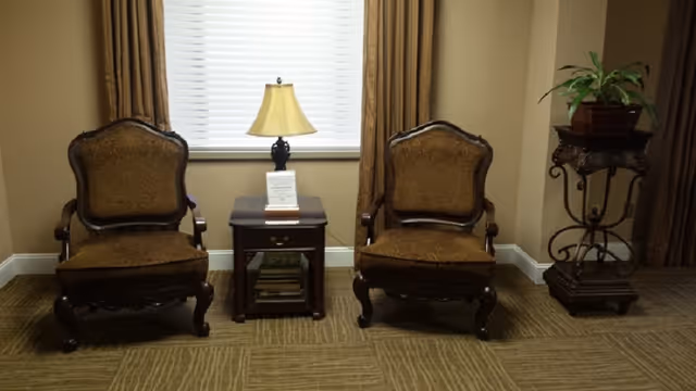Two ornate wooden armchairs with brown upholstery are placed on either side of a small wooden side table with a lamp and a small sign on it. Behind the chairs is a window with closed white blinds and brown curtains. To the right, there is a decorative plant stand with a potted plant on top. The floor is carpeted with a patterned design.
