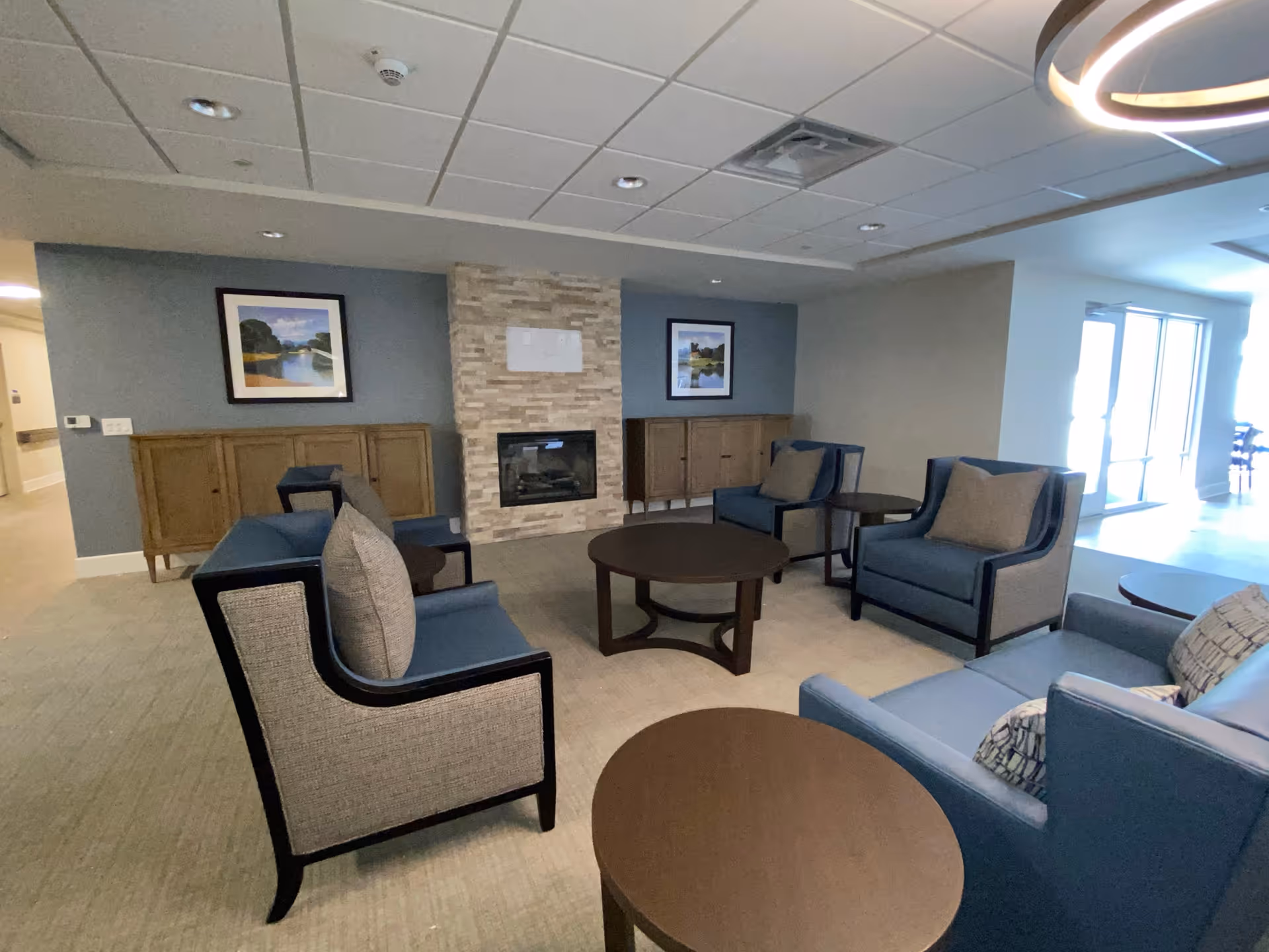 Communal lounge with blue upholstered armchairs, round wooden coffee tables, and a stone fireplace.