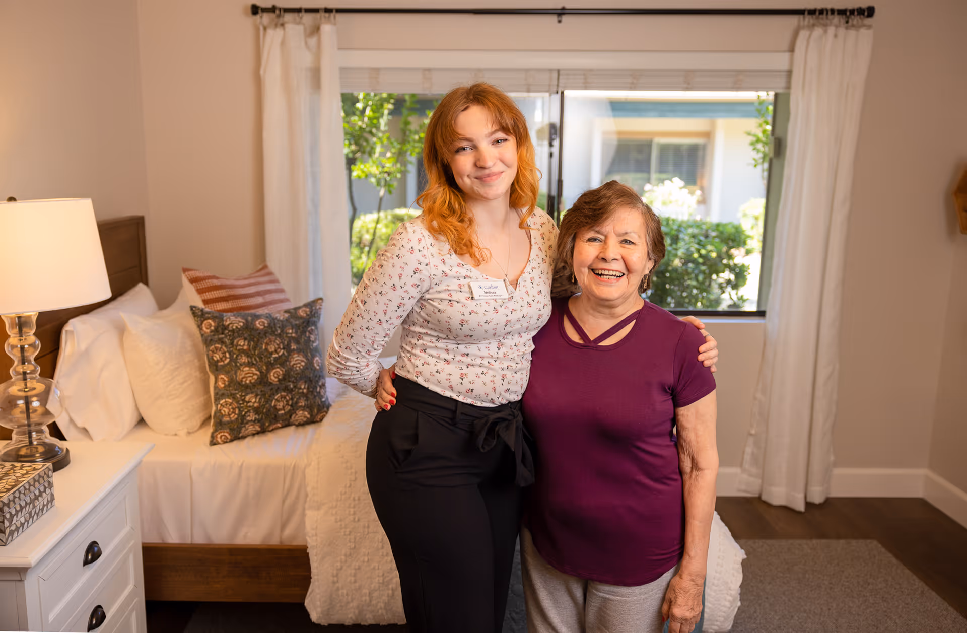 A young woman and an elderly woman standing together and smiling in a bedroom. The bedroom has a bed with white bedding and decorative pillows, a nightstand with a lamp, and a window with white curtains showing greenery outside.