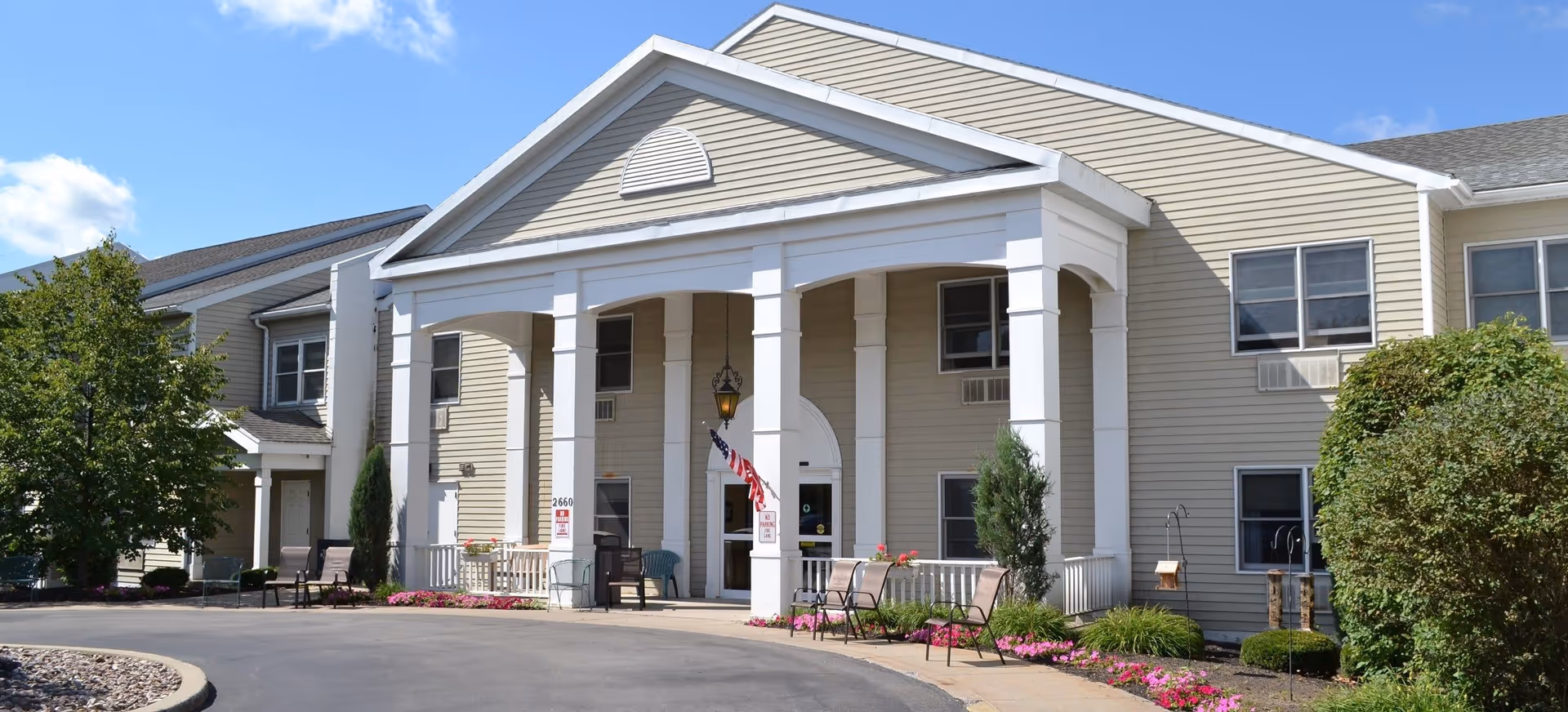 Exterior view of a beige senior living facility building with white columns at the entrance, an American flag hanging near the door, several chairs placed outside, and landscaped flower beds with pink flowers and green bushes under a blue sky with some clouds.