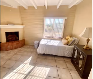 A bright room with a tiled floor featuring a single bed with pillows near a window with vertical blinds. There is a brick fireplace in the corner, a small trash bin next to the bed, and a wooden cabinet with a lamp on top. Sunlight streams through the window, casting shadows on the floor.