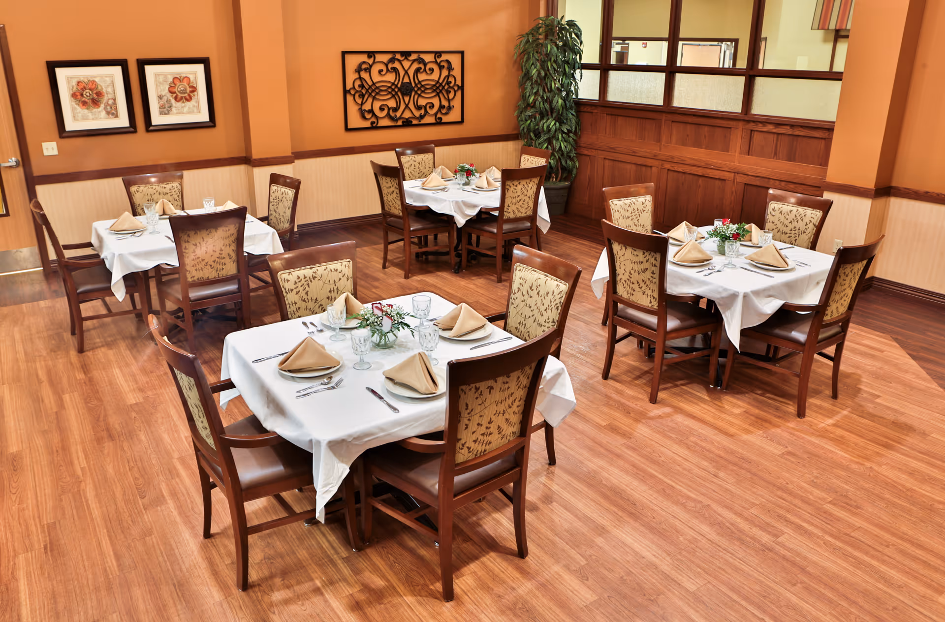A dining room with four square tables covered with white tablecloths, each set with beige folded napkins, glassware, and silverware. The chairs have wooden frames with patterned upholstery. The room has wooden flooring, warm orange walls, decorative wall art, and a tall potted plant in the corner.