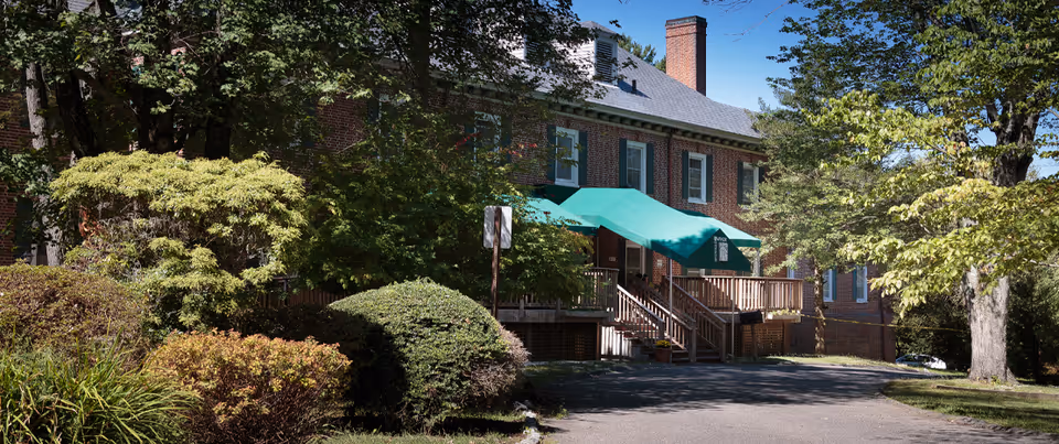 Front exterior of a brick building with green awnings, wooden steps, and surrounding trees and shrubs.