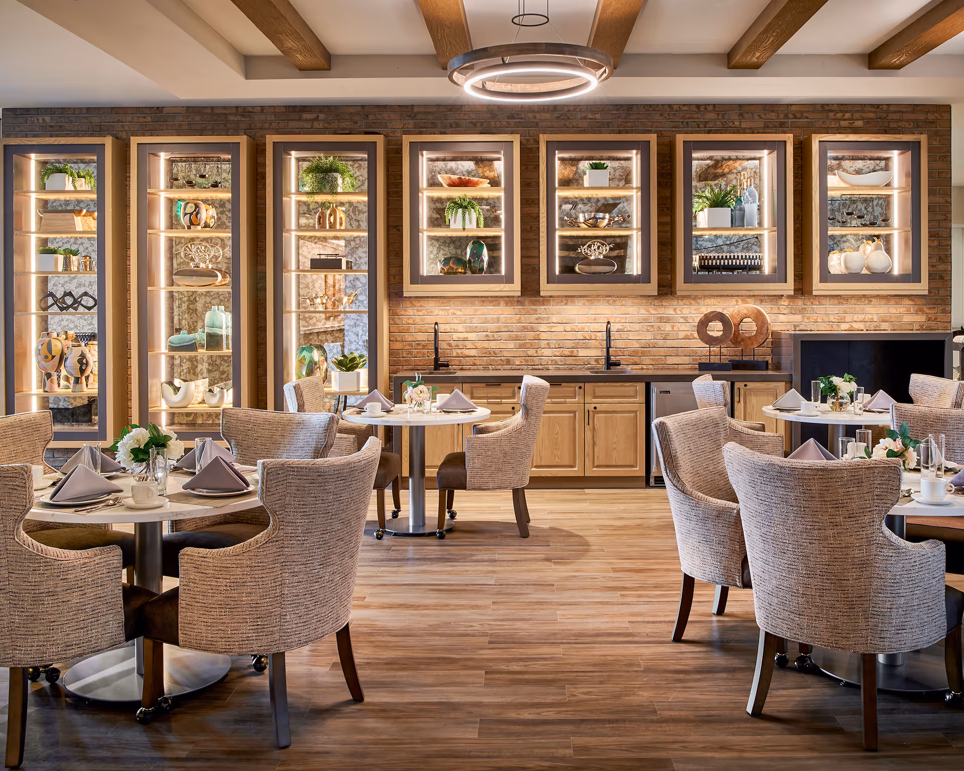 A well-lit dining room with round tables set with napkins, cups, and floral centerpieces. Comfortable upholstered chairs surround the tables. The back wall features wooden cabinets with glass shelves displaying decorative items and plants, set against a brick wall. Wooden beams are visible on the ceiling, and a modern circular light fixture hangs above.