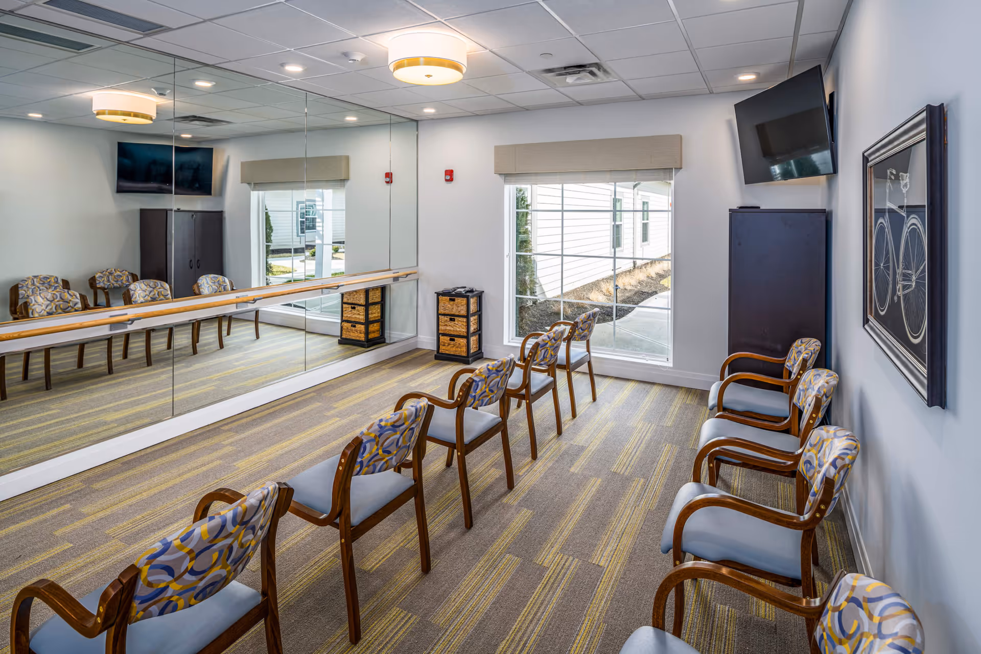 A bright room with a row of wooden chairs with patterned cushions arranged facing a mirrored wall with a ballet barre. The room has a large window with a view outside, a mounted flat-screen TV, a dark cabinet, and a framed picture of a bicycle on the wall. The carpet has a striped pattern in shades of gray and yellow.