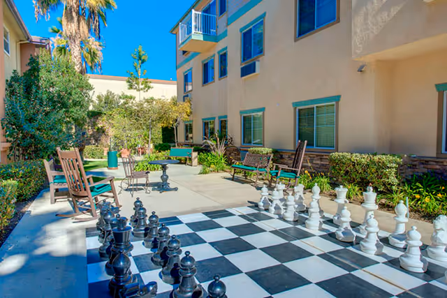 Courtyard patio with a large ground chessboard and oversized chess pieces, benches, and seating beside a multi-story building.