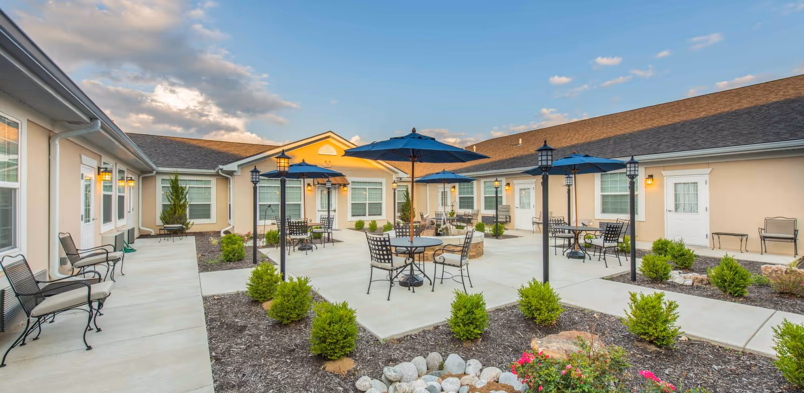 Outdoor courtyard area at Benton House of Lenexa featuring multiple round tables with blue umbrellas, black metal chairs with cushions, lamp posts, and small green shrubs planted in mulched beds. The courtyard is surrounded by a beige building with white-trimmed windows and doors under a partly cloudy sky.