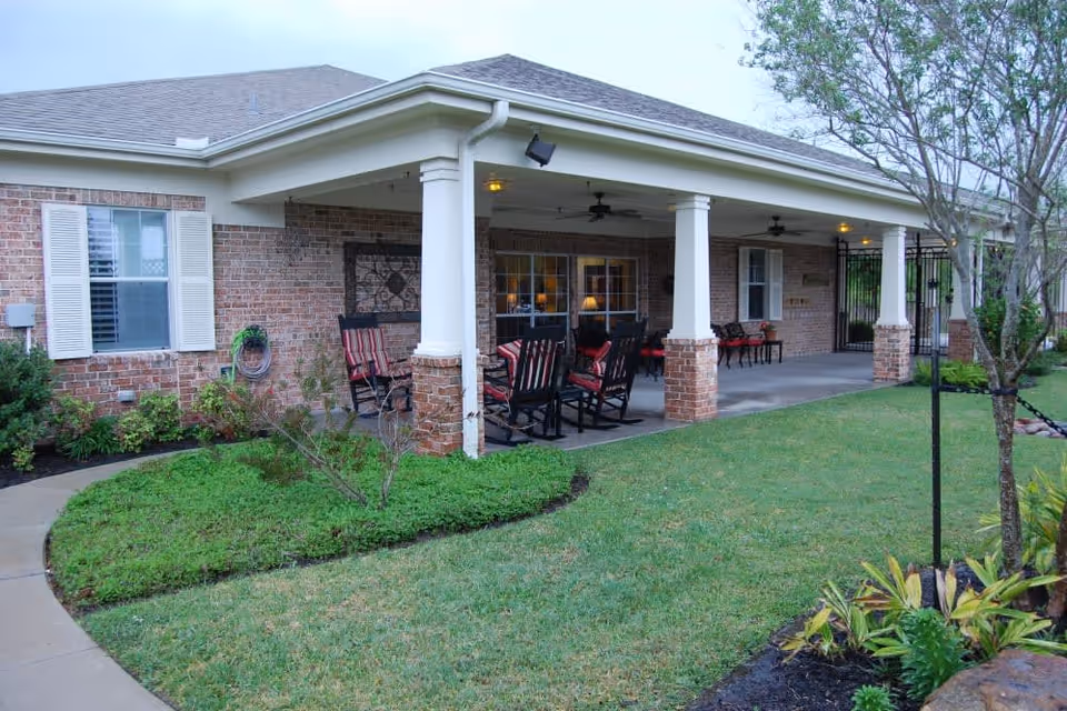 Covered brick patio with rocking chairs and tables outside a single-story assisted living building, bordered by lawn and landscaping.