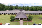 Aerial view of a single-story nursing facility building with a central entrance, surrounded by a well-maintained lawn and trees in the background under a partly cloudy sky.