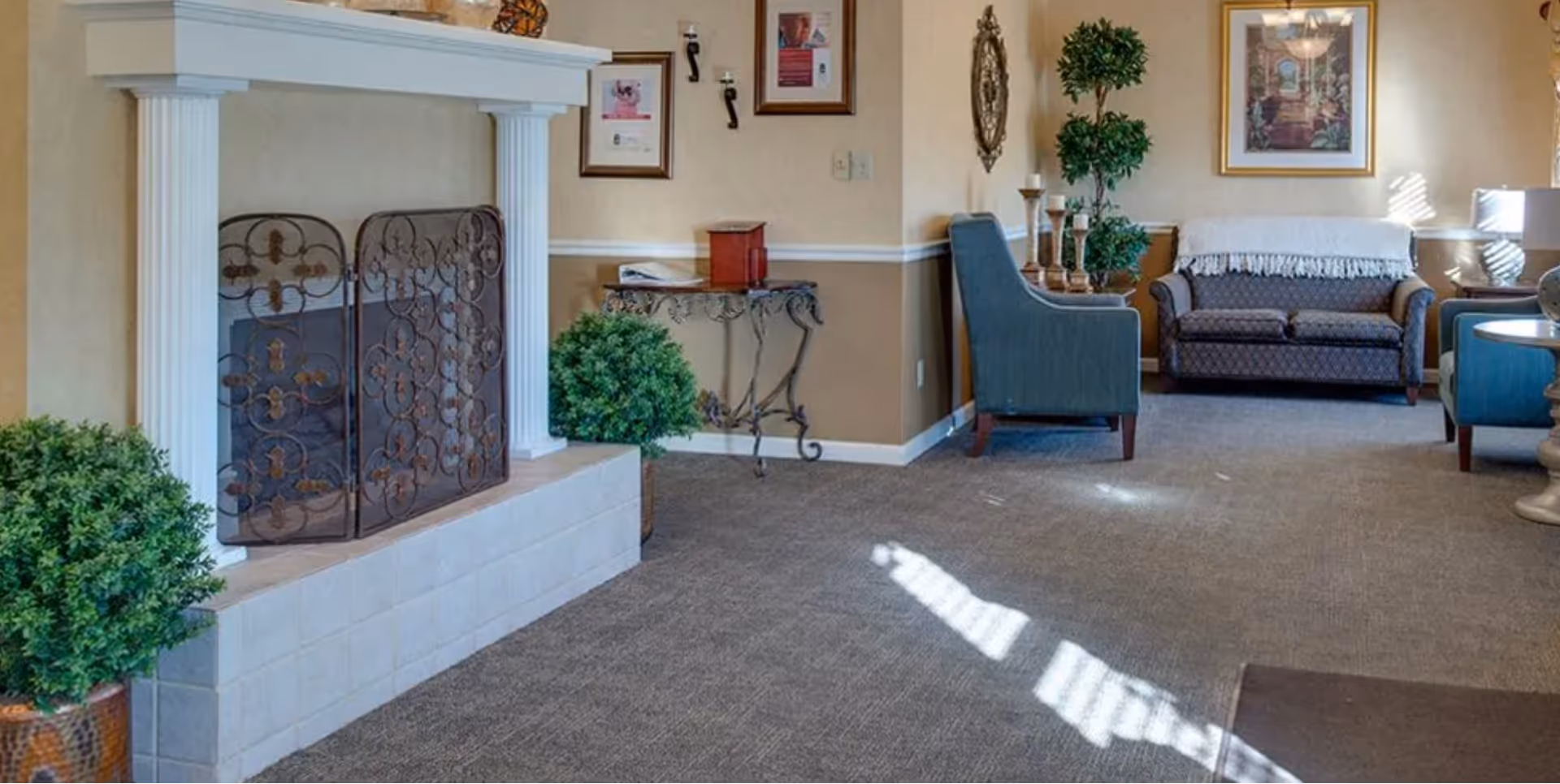 A cozy senior living room area featuring a white fireplace with a decorative metal screen, two green potted plants, a small ornate table with framed pictures on the wall above it, a blue armchair, a patterned sofa with a white throw draped over the back, a side table with a lamp, and a framed painting on the wall. The room has beige walls with white trim and carpeted flooring.