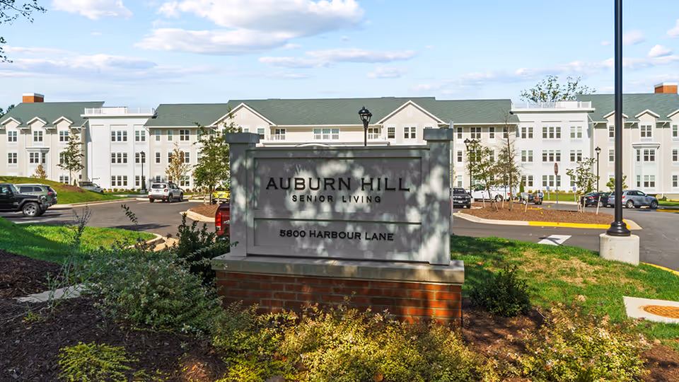 Exterior view of Auburn Hill Senior Living facility with a large sign in the foreground displaying the name and address, surrounded by landscaped greenery and a parking area with cars in front of a multi-story building under a partly cloudy sky.
