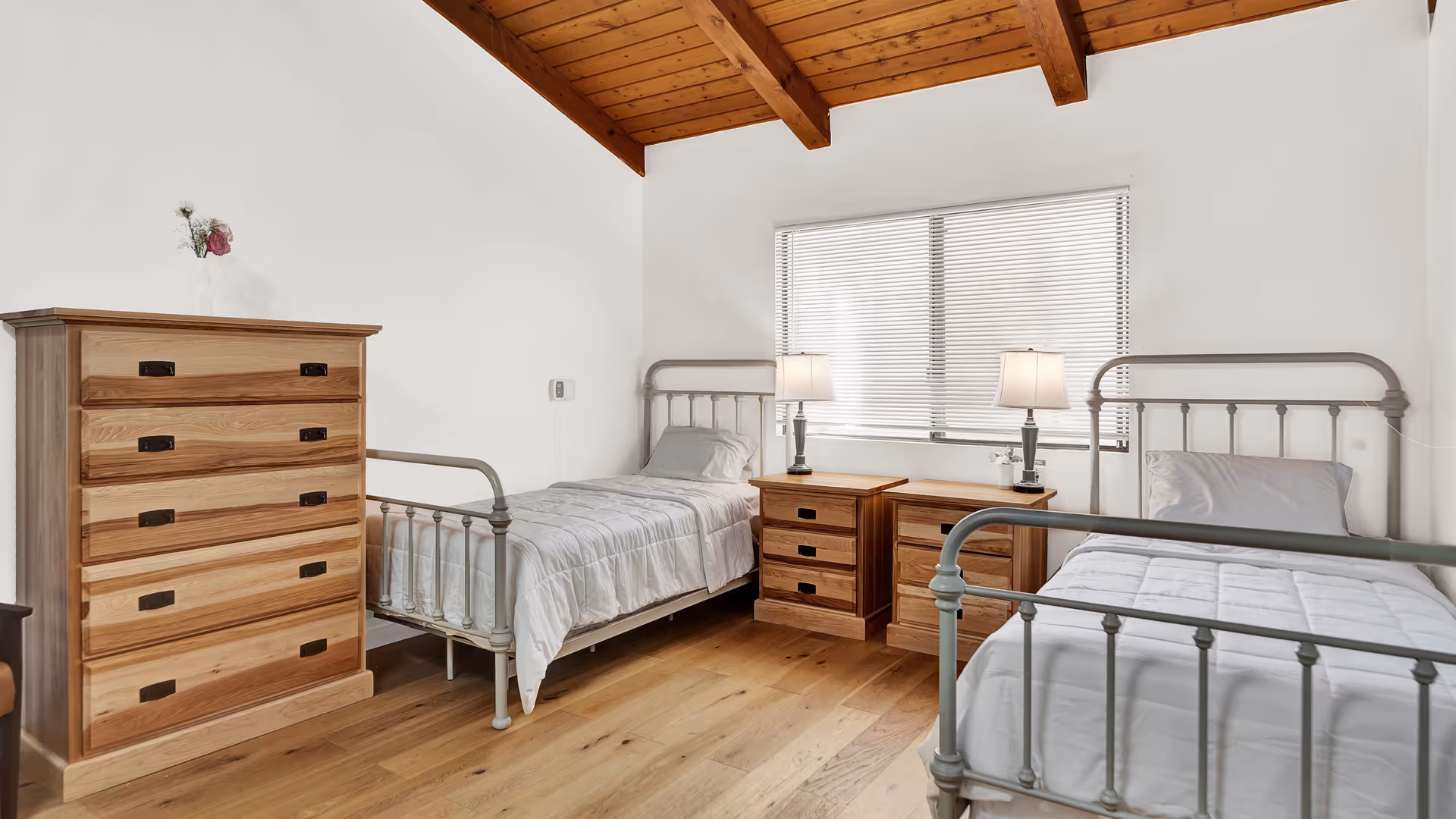 A bedroom with two single metal frame beds, each with white bedding and a pillow. Between the beds are two wooden nightstands with lamps and small decorative items. To the left is a tall wooden dresser with a small vase holding flowers on top. The room has wooden flooring and a wooden ceiling with exposed beams. A window with closed blinds is on the wall behind the beds.