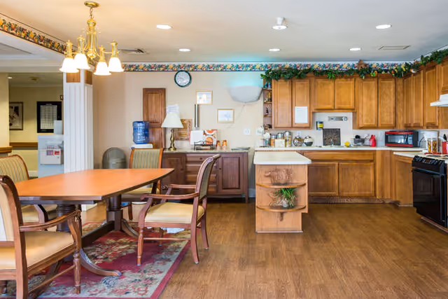 A spacious kitchen and dining area with wooden cabinets, a kitchen island, and a dining table with four chairs. The room has hardwood flooring, a chandelier above the table, a water dispenser, and various kitchen appliances including a microwave and stove. The walls are decorated with a floral border and some framed pictures.