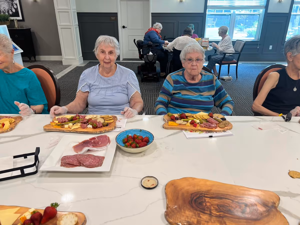 Several elderly women seated at a long table in a communal dining/activity room assembling charcuterie boards and bowls of strawberries.