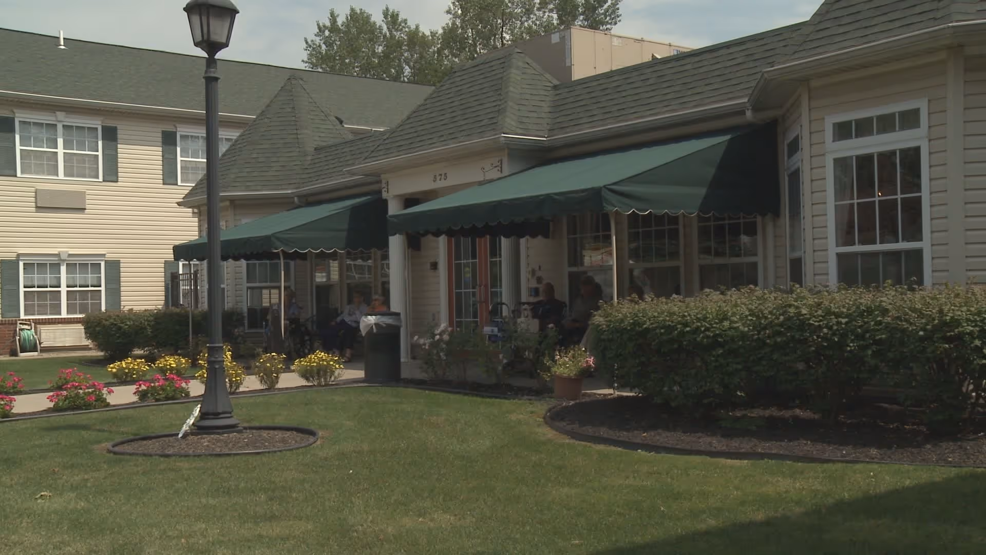 Exterior view of Peregrine Senior Living at Cheektowaga showing a building with green awnings over windows and doors, a well-maintained lawn with flower beds, bushes, and a lamppost in the foreground.